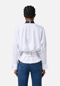 Blusa bianca a manica lunga con colletto nero, caratterizzata da una vita aderente con due dettagli in metallo e una silhouette leggermente oversize.