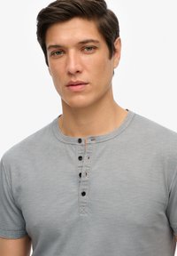 Szara koszulka z krótkim rękawem i okrągłym dekoltem, z zapięciem na guziki i czarnymi guzikami. Miękki, lekki materiał o subtelnej teksturze.