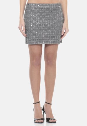 Donna che indossa una minigonna con motivo a maglia d'acciaio argentata e sandali neri con tacco alto e cinturini, in piedi contro uno sfondo semplice.