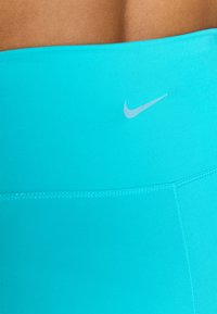 Top sportiv turcoaz cu o textură netedă, având un logo Nike argintiu pe față și detalii cusute de-a lungul cusăturilor.