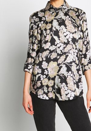 Femme portant un chemisier noir à boutons avec un motif floral rose pâle et vert, manches trois-quarts, accompagné d'un pantalon noir.