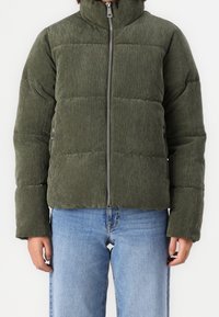 Olivově zelená manšestrová péřová bunda s vysokým límcem, zipovým zapínáním a prošívaným vzorem. Nosená s světle modrými džíny.