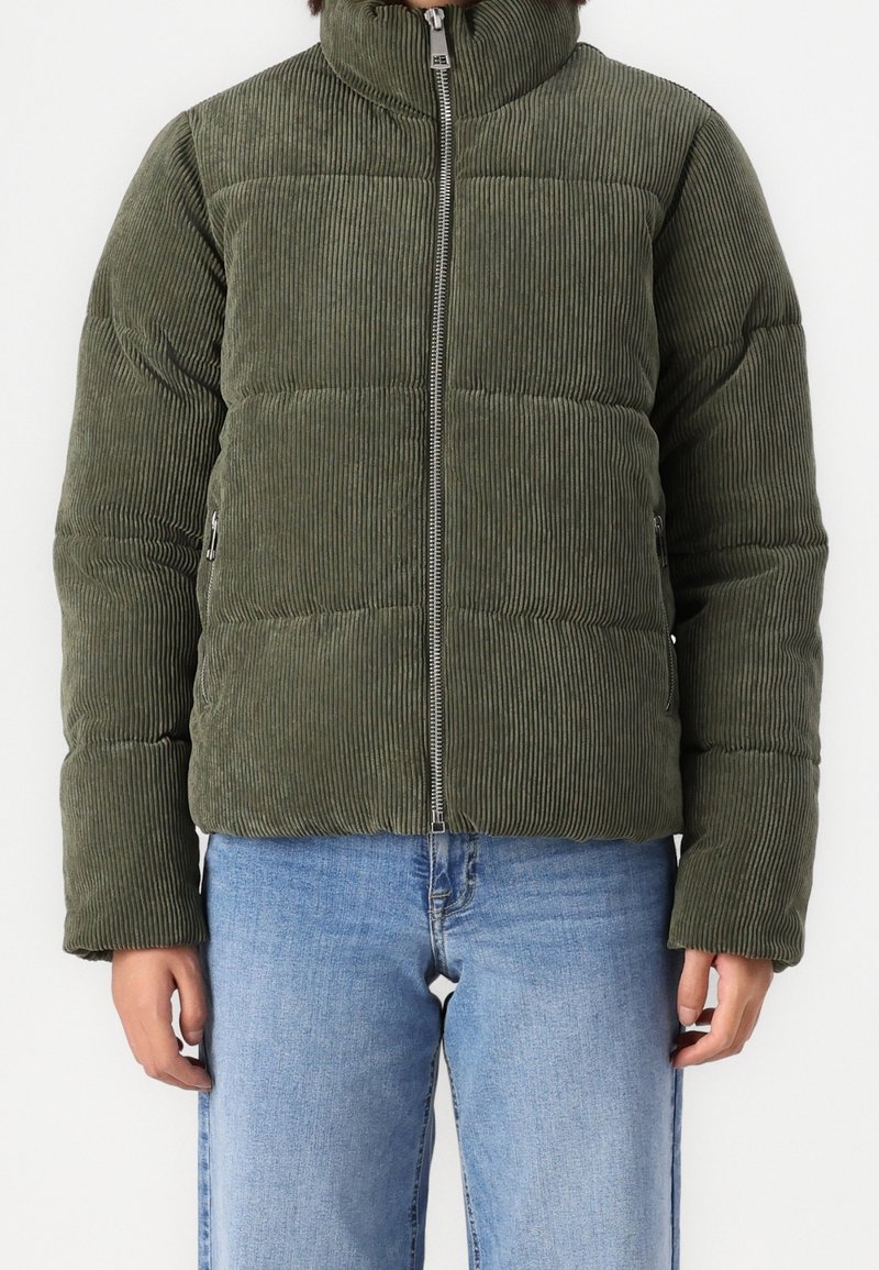 Olivově zelená manšestrová péřová bunda s vysokým límcem, zipovým zapínáním a prošívaným vzorem. Nosená s světle modrými džíny.