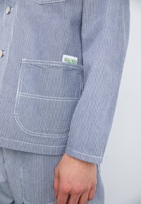 Blue and white striped work jacket made of cotton. Features a chest pocket, white stitching, and a small green "Service Works" tag.