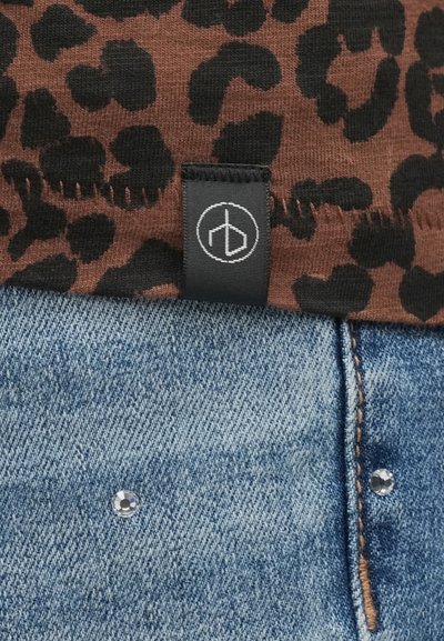 Καφέ ύφασμα με λεοπάρ σχέδιο και μαύρη ετικέτα πάνω από μπλε τζιν ύφασμα με εμφανές πόμολο. Το υλικό φαίνεται απαλό, με λεπτομέρειες ξεφτισμένων άκρων.