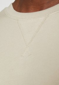 Camisa de manga larga beige con cuello redondo, textura acanalada y detalles de pespunte en el pecho. Acabado de tela suave.
