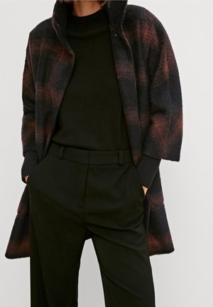 Personne portant un pantalon noir taille haute et un manteau à carreaux foncé avec des teintes brun-rouge sur un pull à col roulé noir.