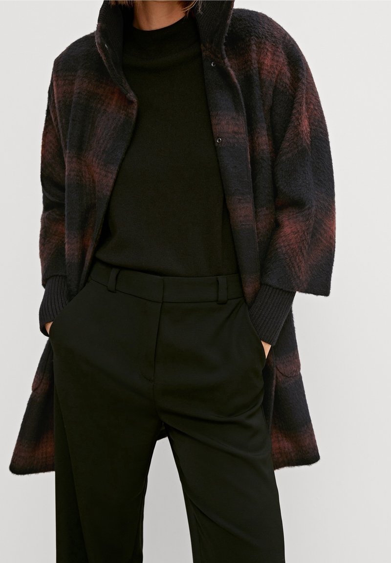 Personne portant un pantalon noir taille haute et un manteau à carreaux foncé avec des teintes brun-rouge sur un pull à col roulé noir.