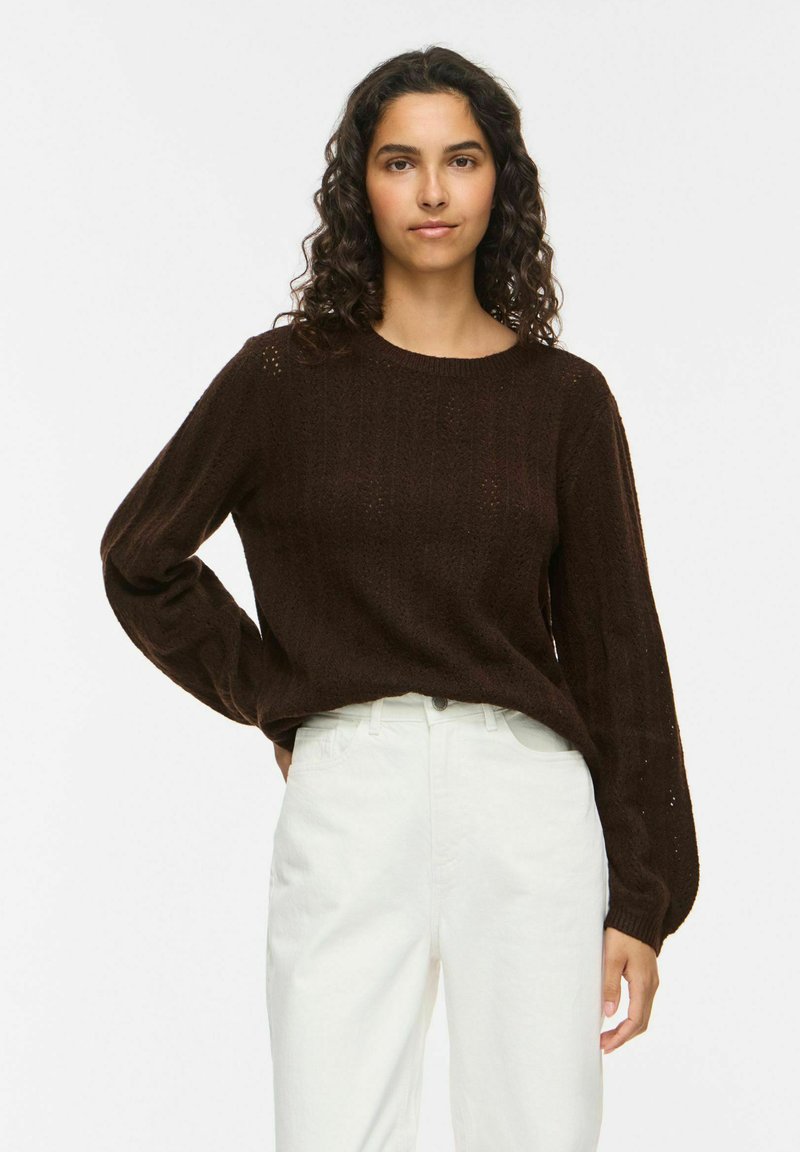 Femme aux cheveux bouclés et foncés portant un pull à manches longues marron foncé et un pantalon blanc, debout avec une main sur la hanche sur un fond uni.