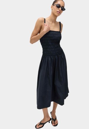 Femme portant une robe midi smockée noire avec bretelles, des sandales à talons noires et des lunettes de soleil noires, debout devant un fond clair uni.