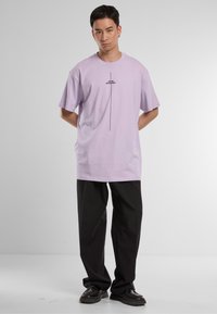 Lavender cotton T-shirt with short sleeves, featuring black text and a vertical line design. Paired with loose black trousers and black shoes.
