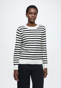 Maglione a righe bianche e nere con scollo rotondo e maniche lunghe, caratterizzato da polsini e orlo a coste. Indossato con pantaloni scuri.