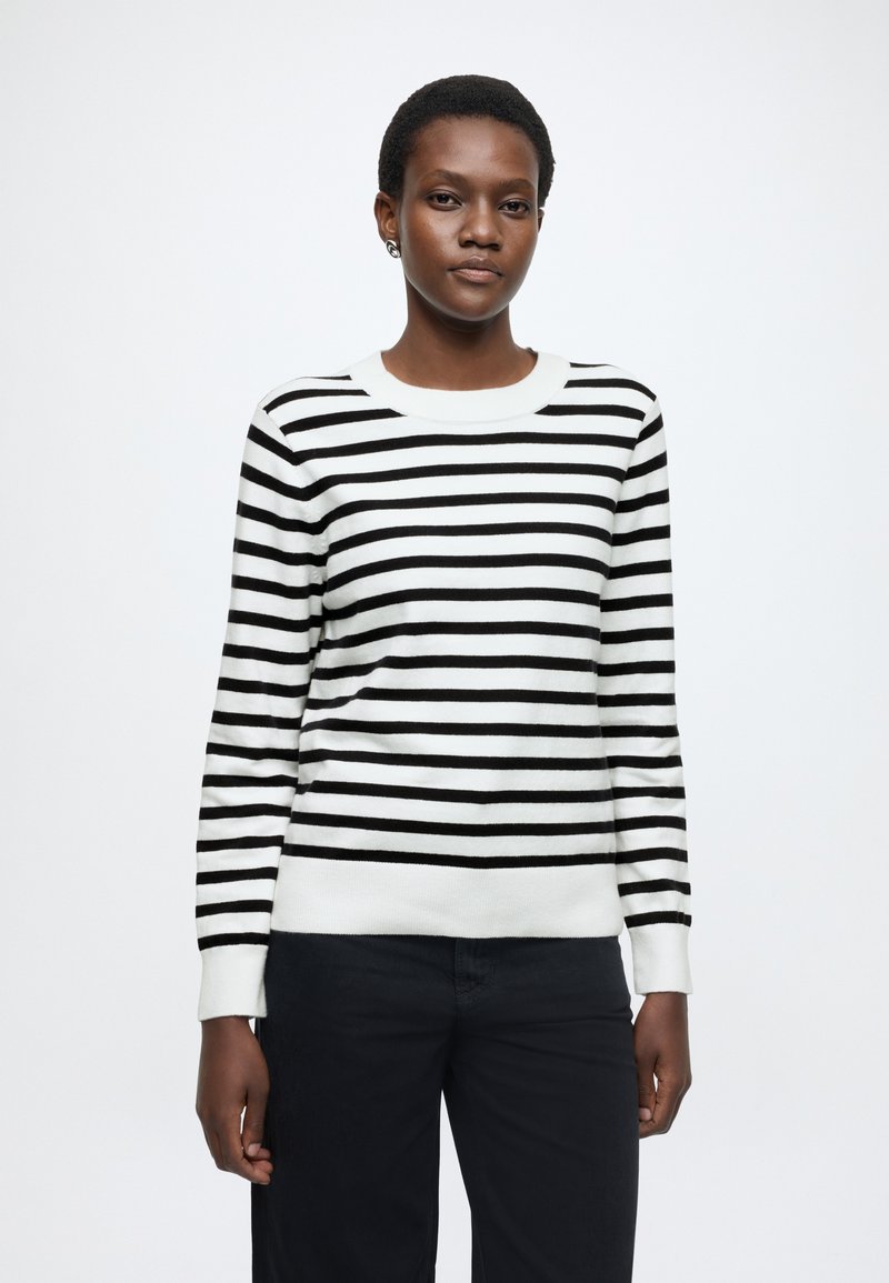 Maglione a righe bianche e nere con scollo rotondo e maniche lunghe, caratterizzato da polsini e orlo a coste. Indossato con pantaloni scuri.