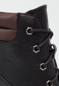 Bottines en cuir noir avec un accent marron au niveau du col, dotées d'œillets hexagonaux, de lacets noirs et d'une surface texturée.