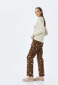 Vaalean beige pitkähihaisessa paidassa, ruskeissa kuvioiduissa housuissa, joissa on jälkiruokamotiiveja, ja pehmeissä beigeissä liukuhihnaisissa tossuissa seisova malli.