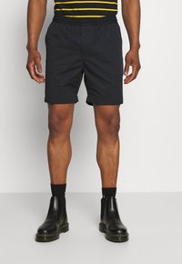 Tmavě modré šortky s elastickým pasem, délka po kolena a hladká textura, spojené s černými kotníkovými botami a tmavými ponožkami.