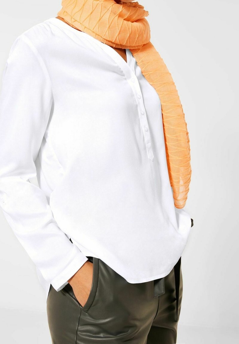 Camicia bianca con bottoni, a maniche lunghe, abbinata a una sciarpa arancione a struttura. La sciarpa è avvolta attorno al collo e presenta un design stropicciato.