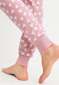 Lichtroze pyjamabroek met witte hartpatronen, voorzien van ribgebreide boorden en een zachte, gladde textuur, gedeeltelijk zichtbaar met blote voeten.