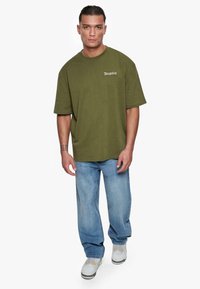 Oliven grønn oversized t-skjorte med frontlogo, kombinert med lyseblå jeans og hvite sneakers med grå detaljer og gule innslag.