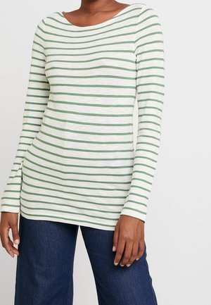 Vrouw draagt een wit shirt met lange mouwen en groene horizontale strepen en een donkere blauwe spijkerbroek, staand tegen een witte achtergrond.