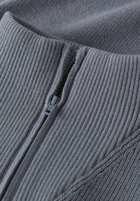 Nahaufnahme eines grauen Strickkleidungsstücks mit einem teilweise geschlossenen Reißverschluss in der Mitte.
