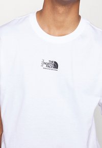 Camiseta blanca de algodón con cuello redondo, que presenta un logo negro "The North Face" y "Never Stop Exploring" impreso en el pecho.