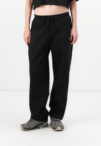 Person iført løse sorte sweatpants med snoretræk i taljen og grå vandresko, stående mod en ensfarvet hvid baggrund.