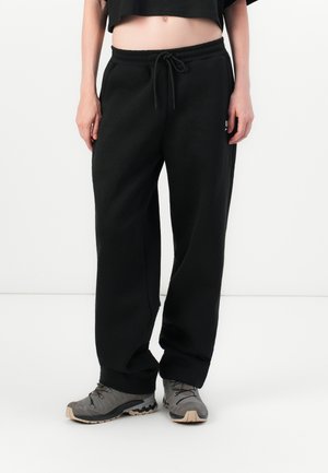 Person iført løse sorte sweatpants med snoretræk i taljen og grå vandresko, stående mod en ensfarvet hvid baggrund.
