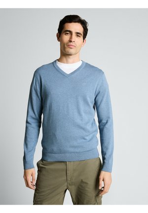 Homme portant un pull col en V bleu clair sur une chemise blanche et un pantalon cargo olive, debout devant un fond uni gris clair.