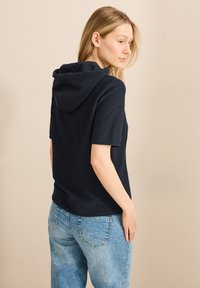 Marineblå, kortärmad hoodie i texturerat tyg. Utrustad med huva, avslappnad passform, kombinerad med ljusblå jeans. Bakvy visas.