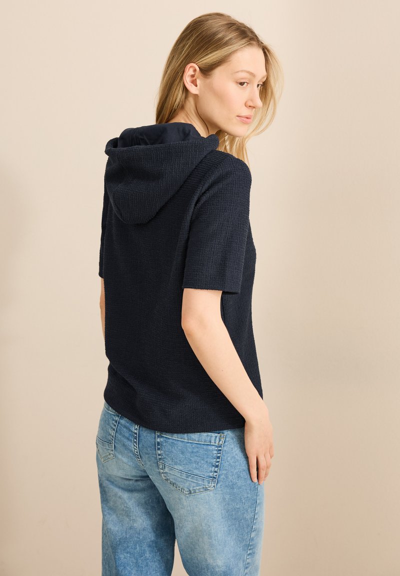 Marineblå, kortärmad hoodie i texturerat tyg. Utrustad med huva, avslappnad passform, kombinerad med ljusblå jeans. Bakvy visas.