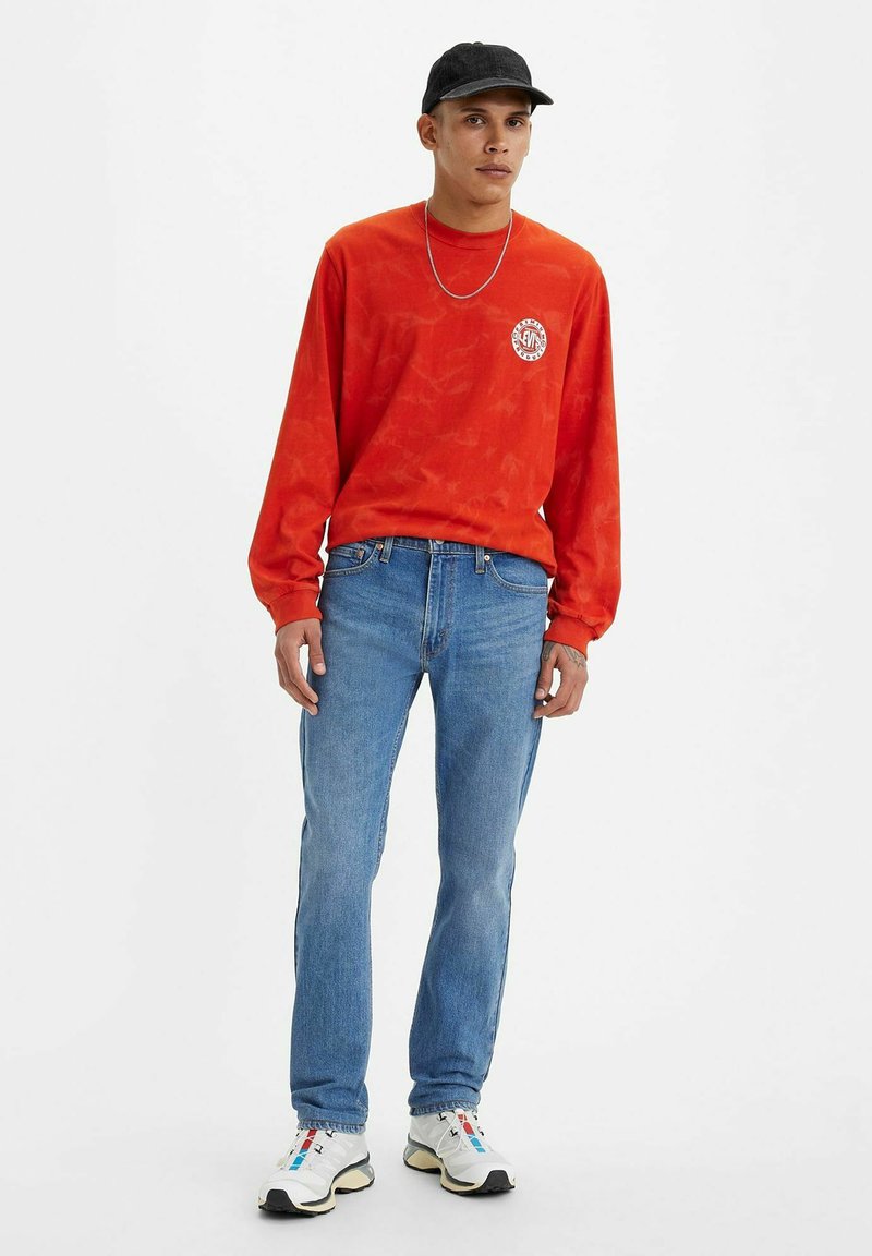 Czerwona bluza z długim rękawem z okrągłym logo, niebieskie dżinsy oraz szare sportowe sneakersy z wielokolorowymi akcentami. Swobodny sportowy wygląd.