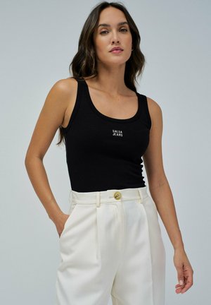 Top de tirantes negro acanalado con escote redondo y logo "Salsa Jeans", combinado con pantalones blancos de cintura alta que presentan un botón dorado.