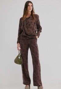 Haut à motif floral marron avec décolleté drapé, associé à un pantalon assorti. Le mannequin tient un sac texturé vert et porte des chaussures à plateforme argentées.