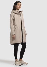 Manteau imperméable beige avec une capuche, des boutons-pression, des poches à l'avant et une coupe droite, associé à des leggings noirs et des baskets beiges.