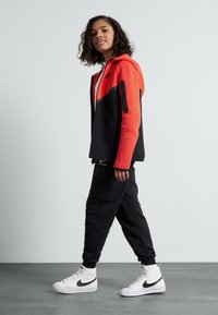 Nike treningsdrakt med rød og svart fargekombinasjon, med en hettejakke med glidelås og smalere bukser, paret med hvite høye sneakers.