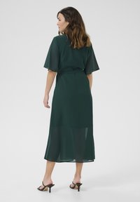 Teal maxi dress with short, sheer sleeves and a tied waist. Features a flowy silhouette with a layered hem. Worn with black heeled sandals.