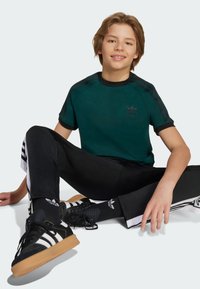 Žalia sportinė marškinėlė su juodais akcentais ir logotipu, derinama su juodomis elastingomis kelnytemis ir juodais sportiniais bateliais su baltais dryžiais ir guminiais padais.