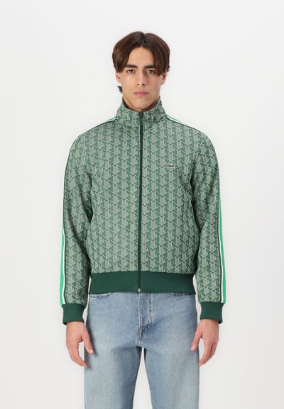 Veste verte à motif avec col haut, fermeture éclair à l'avant et poignets côtelés. Présente des rayures verticales et un petit logo sur la poitrine. Portée par-dessus un denim.