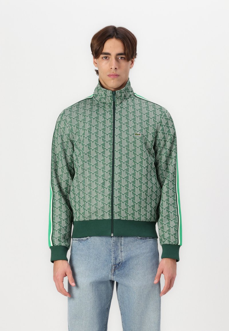 Green patterned jacket with high collar, front zip, and ribbed cuffs. Features vertical stripes and a small logo on the chest. Worn over denim.