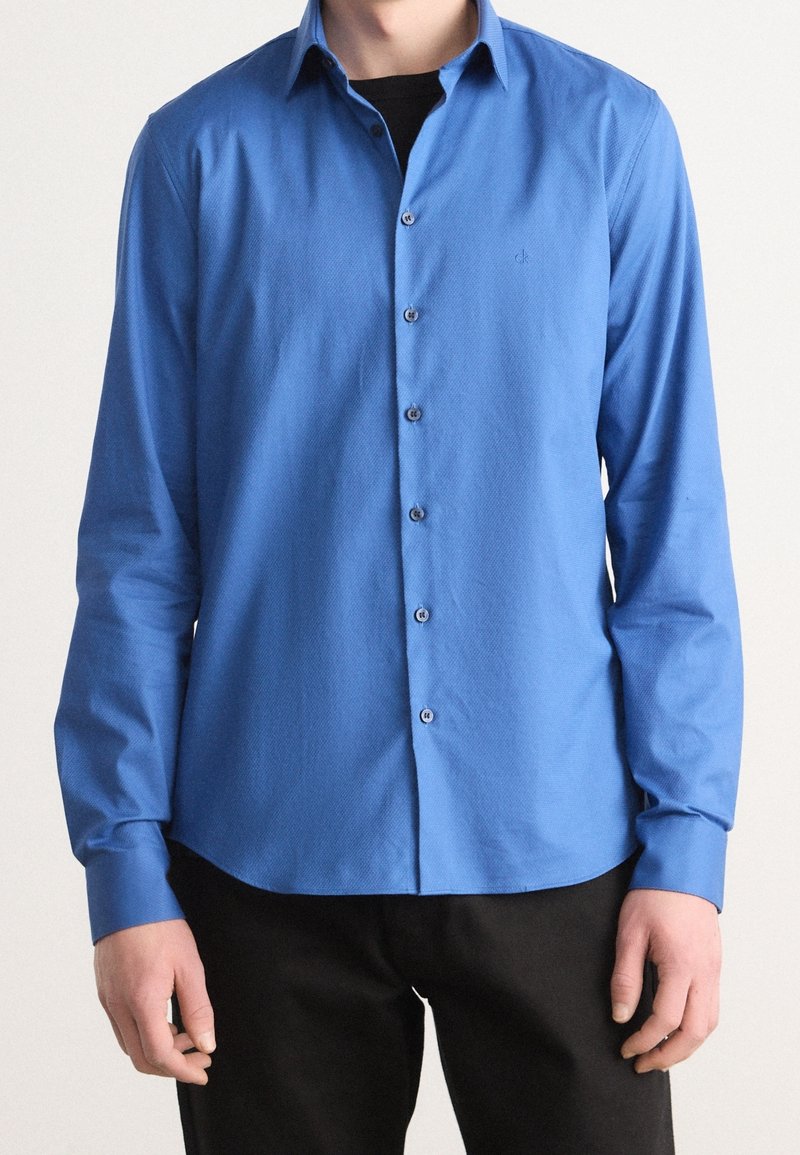 Camicia button-up blu con tessuto testurizzato, maniche lunghe e colletto a punta. Presenta bottoni frontali e un piccolo logo sul petto.