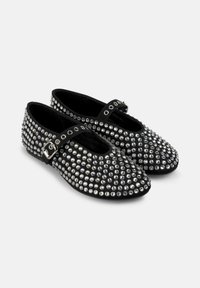 Zapatos planos negros con brillantes tachuelas plateadas que cubren el material. Cuentan con una correa con una hebilla metálica y un diseño de punta redonda.