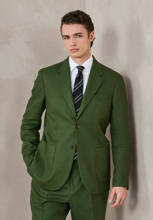 Young man wearing a green suit with two buttons, white shirt, black striped tie, standing against a plain beige wall, hands relaxed.