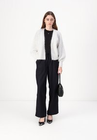 Cardigan blanc en peluche, top noir ajusté, pantalon large à rayures noires, talons noirs et un petit sac à main noir. Textures lisses et couleurs contrastées.