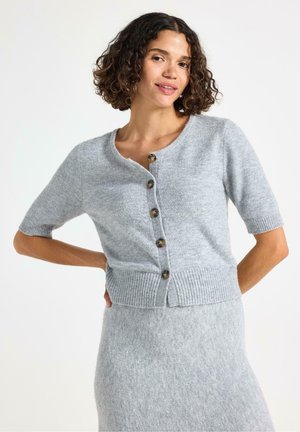 Modella in un cardigan grigio chiaro con maniche corte, che presenta bottoni, sorridente con una mano sul fianco. Lo sfondo bianco semplice mette in risalto l'outfit.