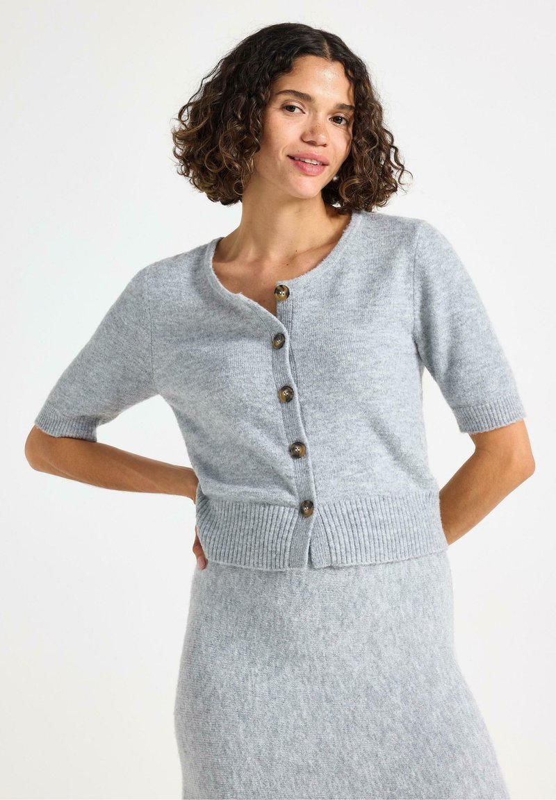 Model i en lys grå cardigan med korte ærmer, der har knapper, smilende med den ene hånd på hoften. Den enkle hvide baggrund fremhæver outfittet.