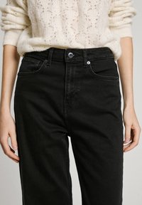 Höga svarta jeans i denim med knäppning, två framfickor och subtila sömnadsdetaljer, ihop med en krämfärgad stickad tröja.