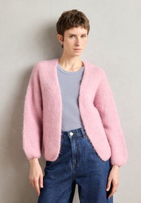 Ružičasti fluffy kardigan sa slobodnim krojem, okruglim izrezom i dugim rukavima, prebačen preko svijetloplavog majice, uparen s trapericama viskog struka.