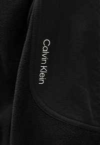 Prim-plan al unui țesătură de fleece negru cu un logo Calvin Klein cusut pe o pată neagră netedă.