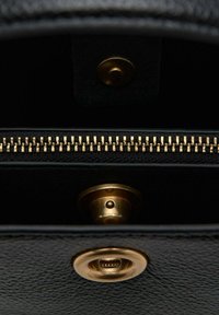 Interiér čiernej koženej tašky, ktorý zobrazuje zlatý zips pozdĺž okraja, kruhový zlatý patentný gombík a hladkú, textúrovanú podšívku.